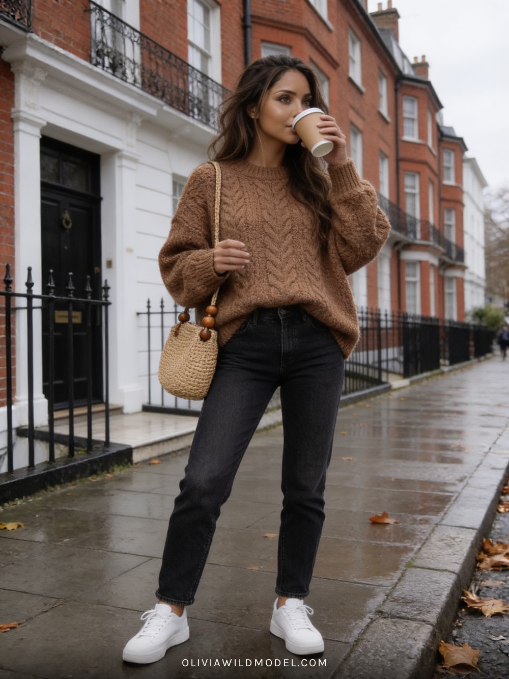 Fall street-style outfit on a London street: oversized coffee-brown chunky cable-knit sweater with dark wash straight jeans, a woven straw shoulder bag and white trainers